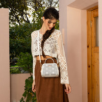 WOMEN IN A IVORY CROCKET CROP TOP AND BROWN SKIRT, HOLDING OUR WHITE GRACIE MINI HANDBAG SET IN A MODERN BACKGROUND. 