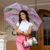 Woman wearing a white crop top and pink shorts smiling while holding the Nicole Lee Violet Blossom Doorman Umbrella and the Nicole Lee Violet Blossom Wide Handbag around her arm against a modern setting. 