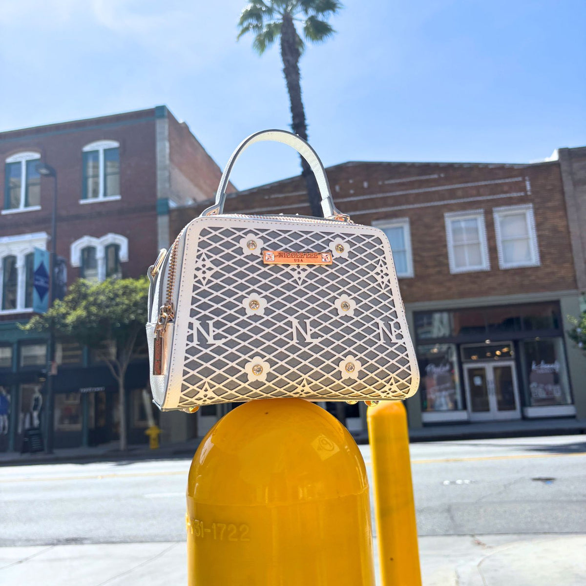 OUR MINI HANDBAG WITH LACER-CUT AND RHINESTONES DETAILS IN COLOR WHITE SET ON A STREET POLE AND BUILDINGS BEHIND.
