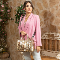 Woman in pink blazer and light blue denim jeans looking to the right while holding a Nicole Lee Calista handbag in metallic gold with gold-tone hardware details. The woman is carrying it by the top handles while also wearing the logo webbed shoulder strap to show varied carrying options and versatility. Set against a modern, rustic background with a bench and flowers hanging behind her. 