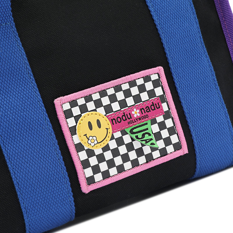 Close up detail of the Rowan handbag in black by Nodu Nadu. Adorned with an embroidered checker-print stitch label bordered in baby pink with a yellow smiley face and flower and a nodu nadu hollywood label with a green USA flag.  Bold blue straps and royal purple trim add pops of color against the timeless black colorway.