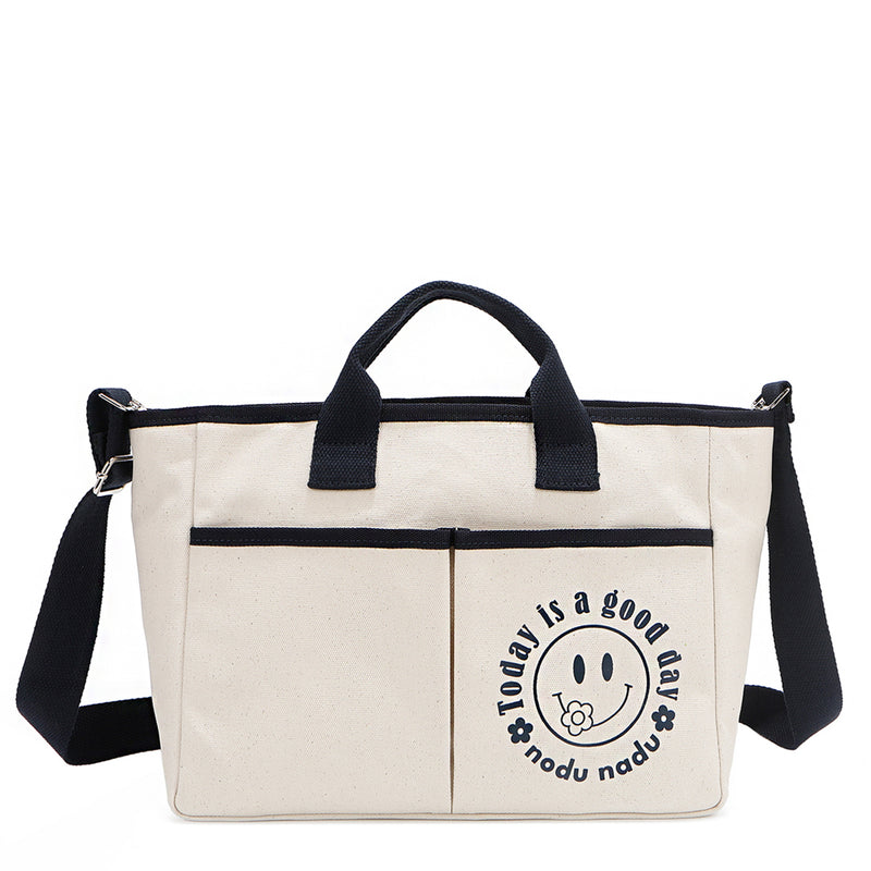 Back angle of the Emery handbag by Nodu Nadu in classic canvas-beige with navy blue trim. Made of polyester with dual top handles and a convenient black slip pocket to keep most-used items like your phone, keys, and planner close at hand. Dual top handles with a 3" drop and an optional, adjustable shoulder strap allow you to carry this tote by the top handles as a handbag or across the body as a crossbody or shoulder bag! The options are endless to mix and match according to your style needs. 