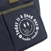 Close up detail of the Emery tote bag in navy blue with moss green trim and straps and a white "today is a good day" quote wrapped around a white smiley face with a cute little flower on its smile. Simple, minimalist, yet positive and on-trend, spread the good vibes with this classic statement. 