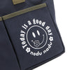 Close up detail of the Emery tote bag in navy blue with moss green trim and straps and a white "today is a good day" quote wrapped around a white smiley face with a cute little flower on its smile. Simple, minimalist, yet positive and on-trend, spread the good vibes with this classic statement. 