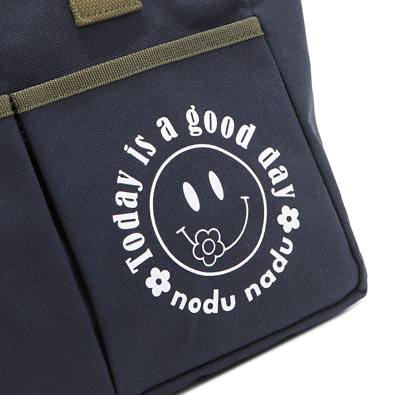Close up detail of the Emery tote bag in navy blue with moss green trim and straps and a white "today is a good day" quote wrapped around a white smiley face with a cute little flower on its smile. Simple, minimalist, yet positive and on-trend, spread the good vibes with this classic statement. 