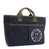 With sleek trim details and a feel-good message, this everyday tote is here to remind you that yes, today (and every day) is a good one. Designed for both style and function, it features plenty of compartments like 2 front slip pockets. Front angle pictured in classic navy blue with moss green trim and straps. White smiley face adorns the front with a cute little flower on its smile in this rectangular silhouette. 