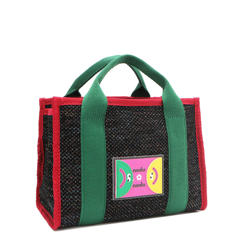Front angle view of the Nodu Nadu small Marley tote bag in black.  Featuring dual top handles with a 3.5" drop and adorned with a logo stitched nodu nadu patch with a smiley face and sad face. Metallic shimmering black canvas with forest green strap accents complimented with a bold red trim. 