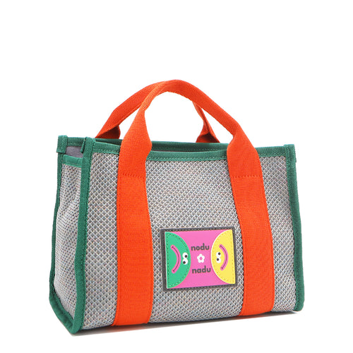Front angle view of the Nodu Nadu small Marley tote bag in gray.  Featuring dual top handles with a 3.5" drop and adorned with a logo stitched nodu nadu patch with a smiley face and sad face. Metallic shimmering gray canvas with bold orange strap accents complimented with a green trim. 