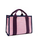 Back angle view of the Nodu Nadu small canvas Marley tote Bag in pink. Accented with eggplant purple straps and a navy blue trim, this canvas features a subtle metallic shimmer that hits the light whenever you walk for a head-turning detail. 