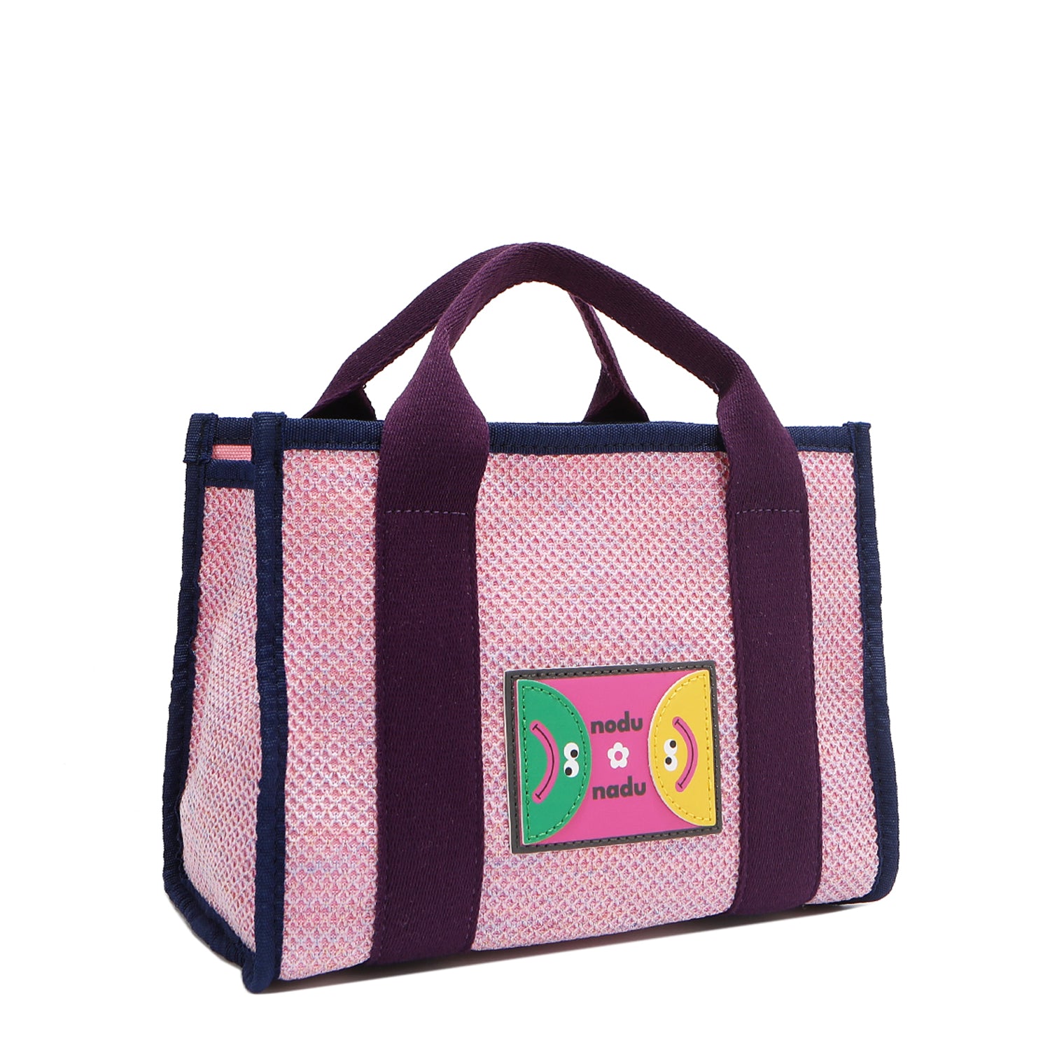 Front angle view of the Nodu Nadu small Marley tote bag in pink.  Featuring dual top handles with a 3.5" drop and adorned with a logo stitched nodu nadu patch with a smiley face and sad face. Metallic shimmering light pink canvas with eggplant purple strap accents complimented with a navy blue trim. 
