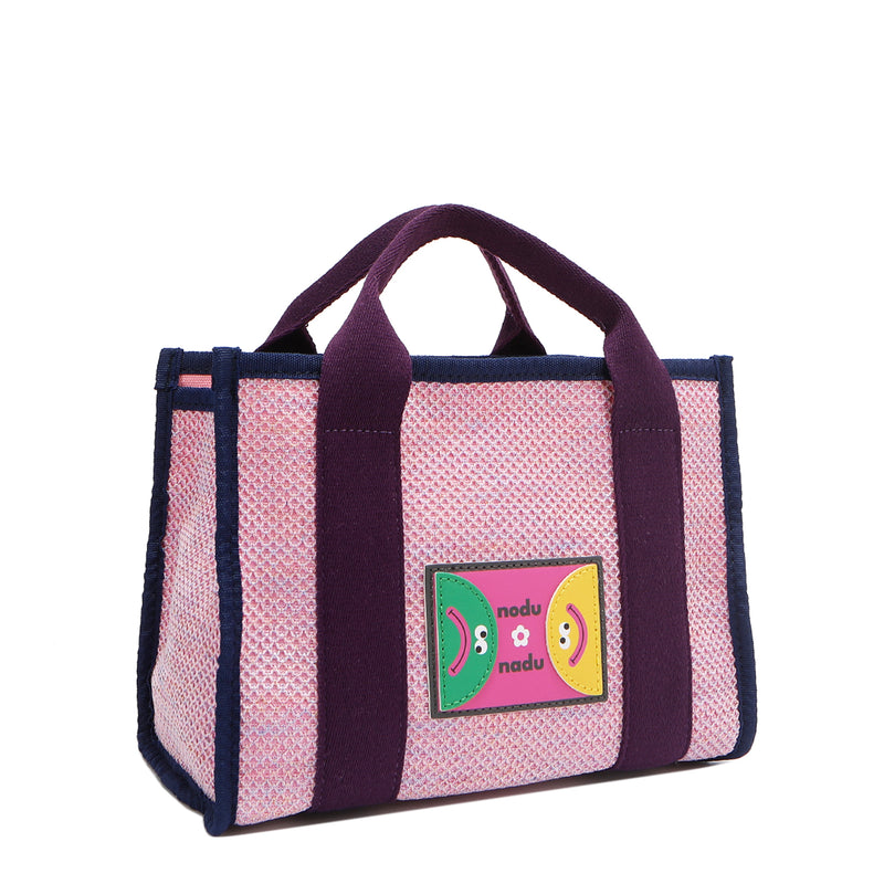 Front angle view of the Nodu Nadu small Marley tote bag in pink.  Featuring dual top handles with a 3.5" drop and adorned with a logo stitched nodu nadu patch with a smiley face and sad face. Metallic shimmering light pink canvas with eggplant purple strap accents complimented with a navy blue trim. 