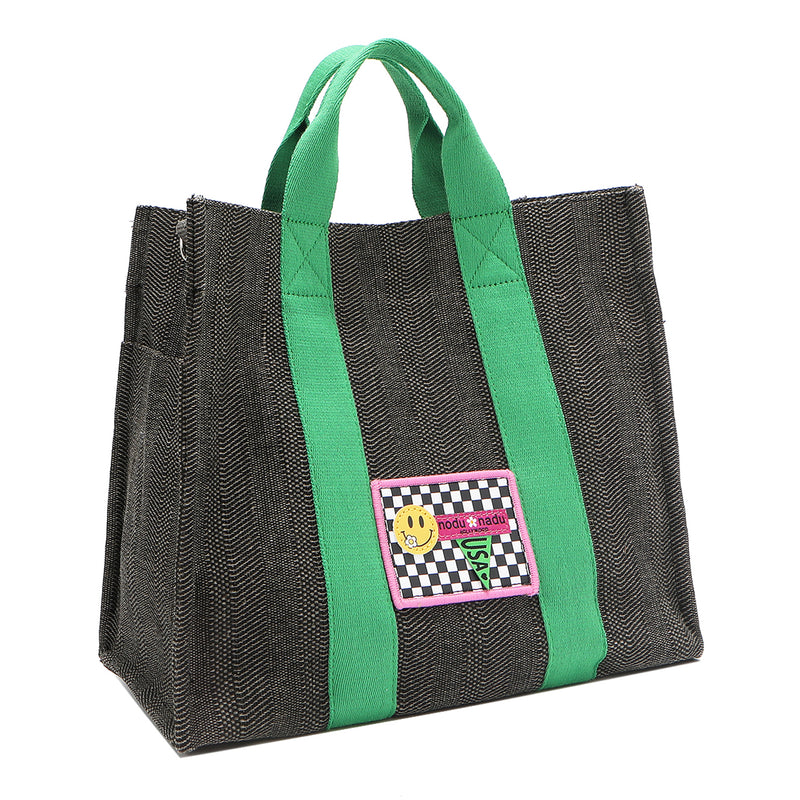 Front angle view of the Nodu Nadu Sawyer Bag in Black with grass green strap accents. Charcoal black canvas adorned with a stitched logo patch in black and white checker print with nodu nadu smiley face. Front angle shows the side slip pocket to store essentials that you want close within reach like your phone, keys, wallet, lipstick, and more!