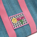 Close-up macro detail shot of the Nodu Nadu Sawyer Bag in Blue. Baby pink strap accents adorn this canvas everyday bag. The front is adorned with an embroidered stitch logo patch in a black and white checker print with a nodu nadu smiley face and daisy flower on its smile. Nodu Nadu tag made in hollywood with a green USA flag to showcase our proud origins. 