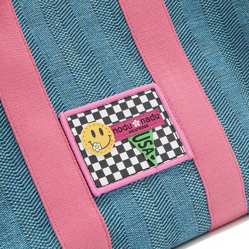 Close-up macro detail shot of the Nodu Nadu Sawyer Bag in Blue. Baby pink strap accents adorn this canvas everyday bag. The front is adorned with an embroidered stitch logo patch in a black and white checker print with a nodu nadu smiley face and daisy flower on its smile. Nodu Nadu tag made in hollywood with a green USA flag to showcase our proud origins. 