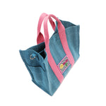Top angle of the Nodu Nadu Sawyer Bag in blue with baby pink dual handles and a checker print stitched logo with a smiley face. Side D-ring for optional shoulder strap attachment and side slip pocket to store everyday must-haves like makeup, wallet, keys, and more conveniently within reach! 
