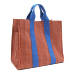 Back angle view of the Nodu Nadu Sawyer Bag in Orange/Sienna brown with bold blue accents. Rectangular side view of the canvas bag shows the side slip pocket that can store any essentials you want close at hand. 