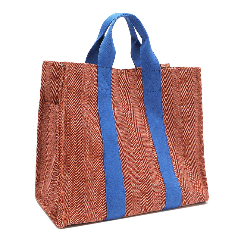 Back angle view of the Nodu Nadu Sawyer Bag in Orange/Sienna brown with bold blue accents. Rectangular side view of the canvas bag shows the side slip pocket that can store any essentials you want close at hand. 