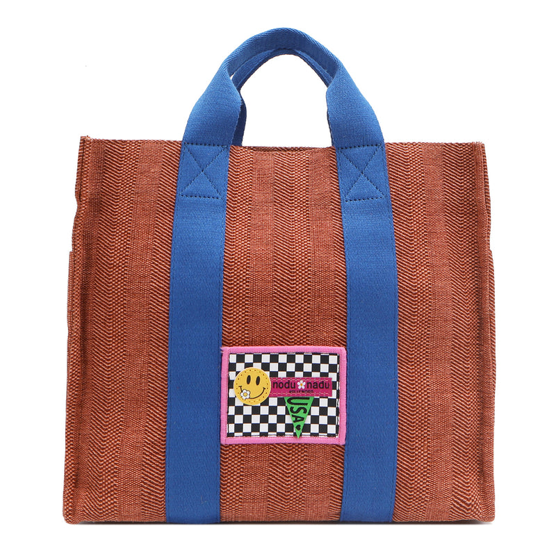 Your must-have carryall for school, weekends, and everything in between! Designed with a soft, relaxed silhouette, the Nodu Nadu Sawyer in Orange offers plenty of space to keep notebooks and loose essentials effortlessly organized—so you're always ready to grab and go! Burnt orange/sienna brown-colored canvas paired with bold blue strap accents. 