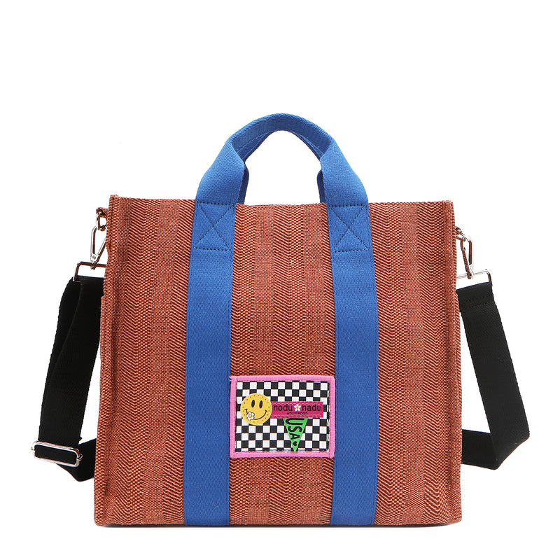 Nodu Nadu Sawyer Bag in Orange/Sienna Brown with bold blue strap accents and paired with an adjustable, optional black wide shoulder strap. Wear this bag by the top handles or across the body thanks to the versatile shoulder strap options. 