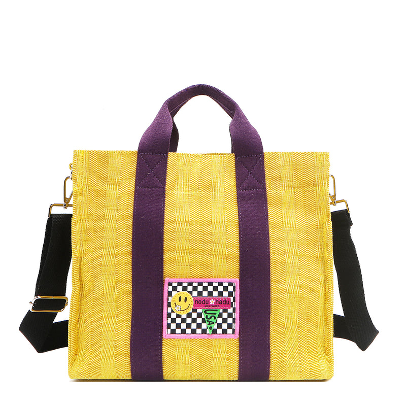 Nodu Nadu Sawyer Bag in Yellow with eggplant purple strap accents and paired with an adjustable, optional black wide shoulder strap. Wear this bag by the top handles or across the body thanks to the versatile shoulder strap options. 