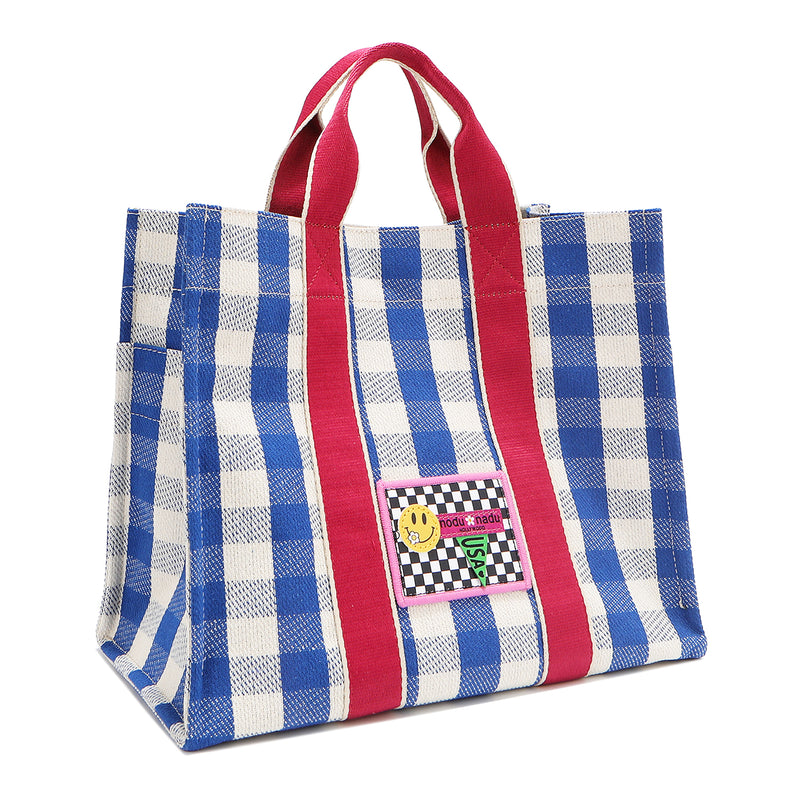 Stylish Nodu Nadu Blair Bag captured at a front angle, showing depth, structure, and playful design details. Pictured in the color blue with blue and white gingham print complimented with hot pink webbed dual straps. Slightly tilted front angle shows the side slip pocket detail. 