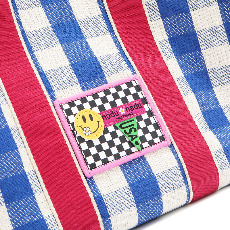 Close-up macro shot of the Nodu Nadu Blair Bag in blue with blue and white gingham print and hot pink shoulder strap accents. The front is adorned with an embroidered checker print stitch logo with baby pink trim and a smiley face and nodu nadu logo with USA flag.